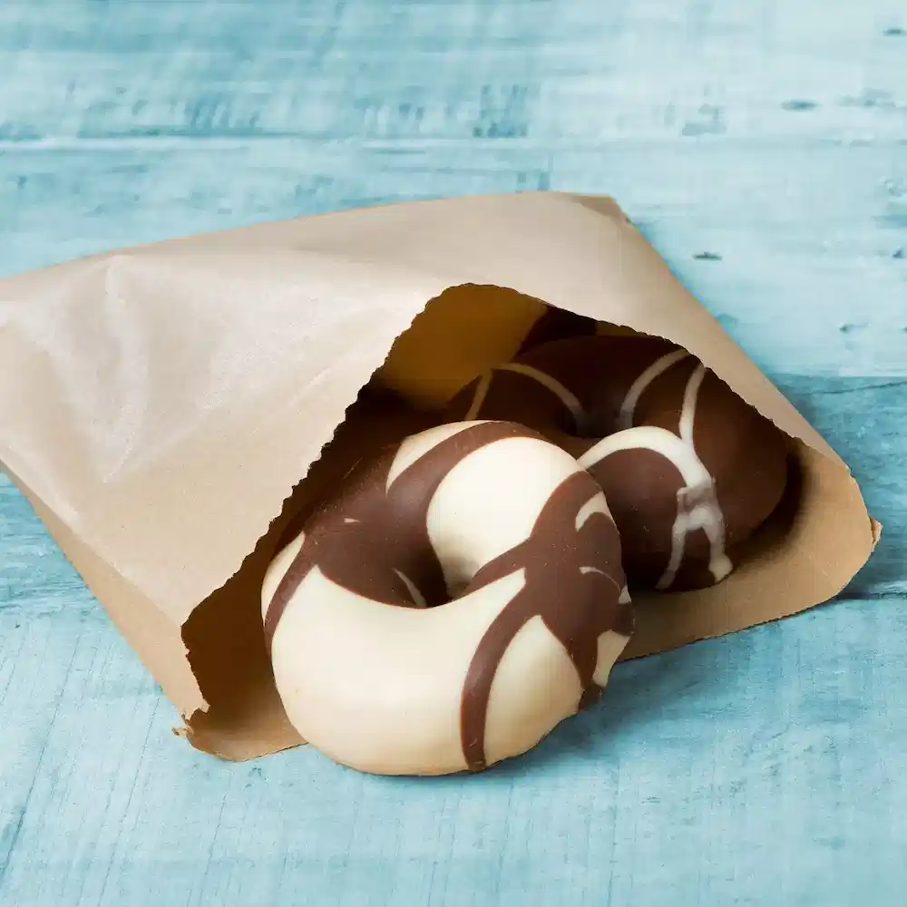Chocolade-glazuurde donuts, gedeeltelijk zichtbaar in een bruine papieren zak op een blauwe houten ondergrond.