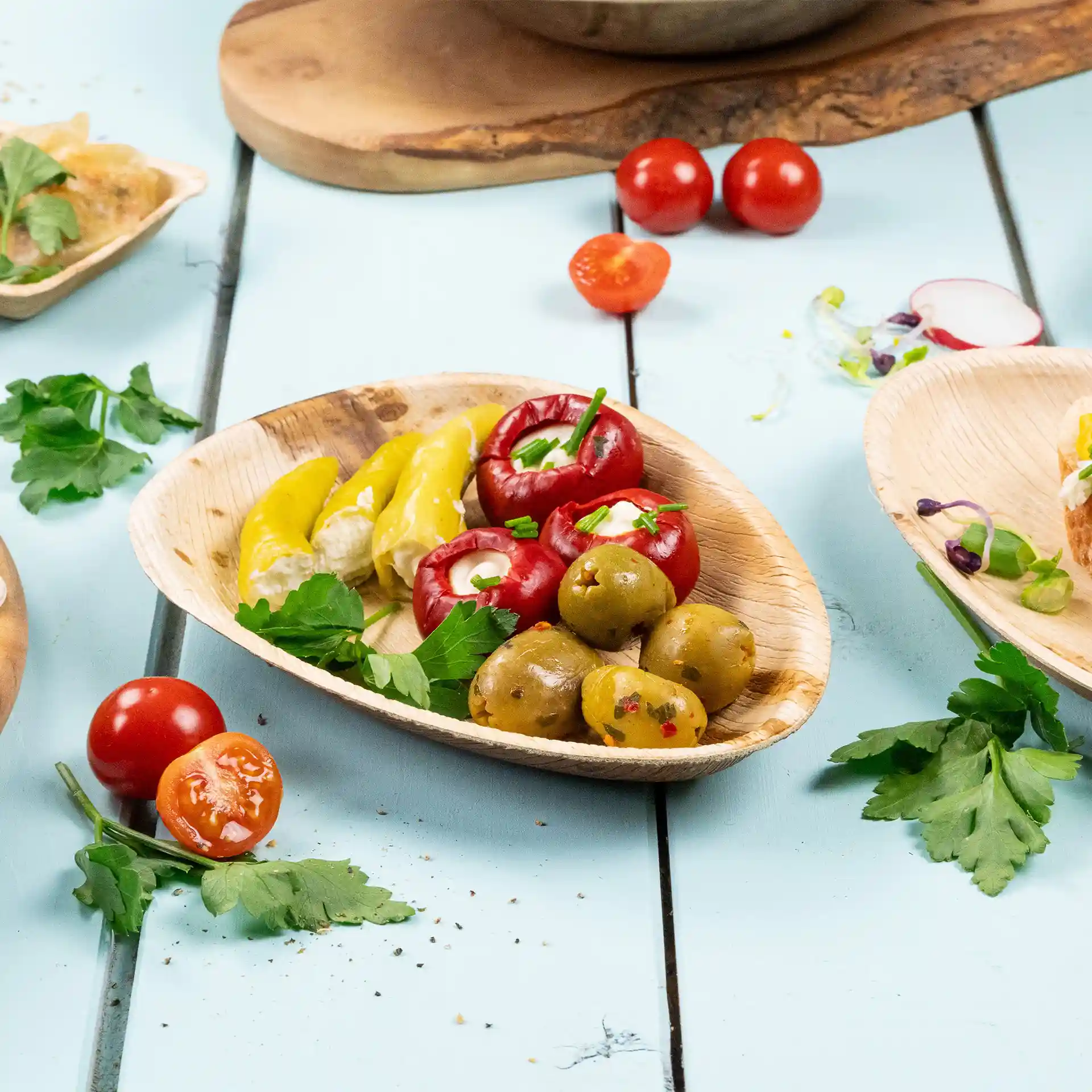 Ovale palmbladplaat met gele paprika, rode gevulde tomaten, groene olijven en peterselie op een blauwe ondergrond.