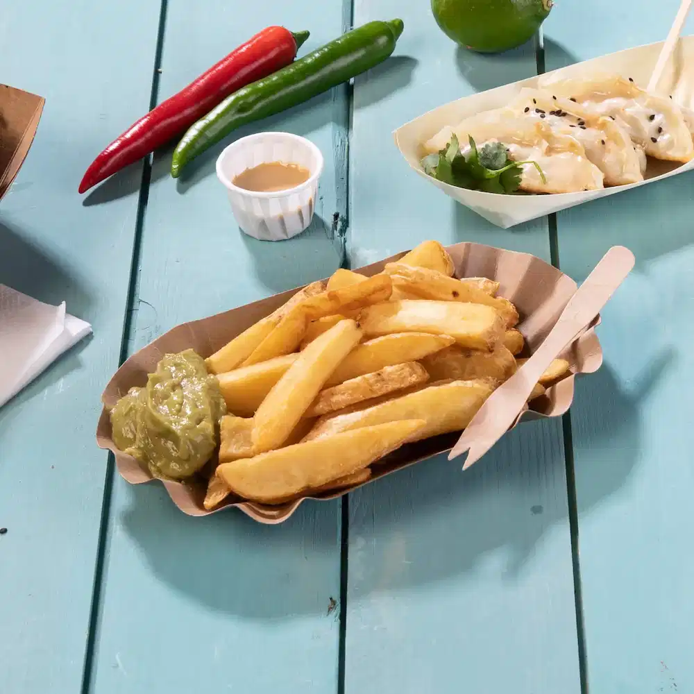 Mand met frietjes, groene saus, houten bestek en chilipepers op een blauwe houten tafel.