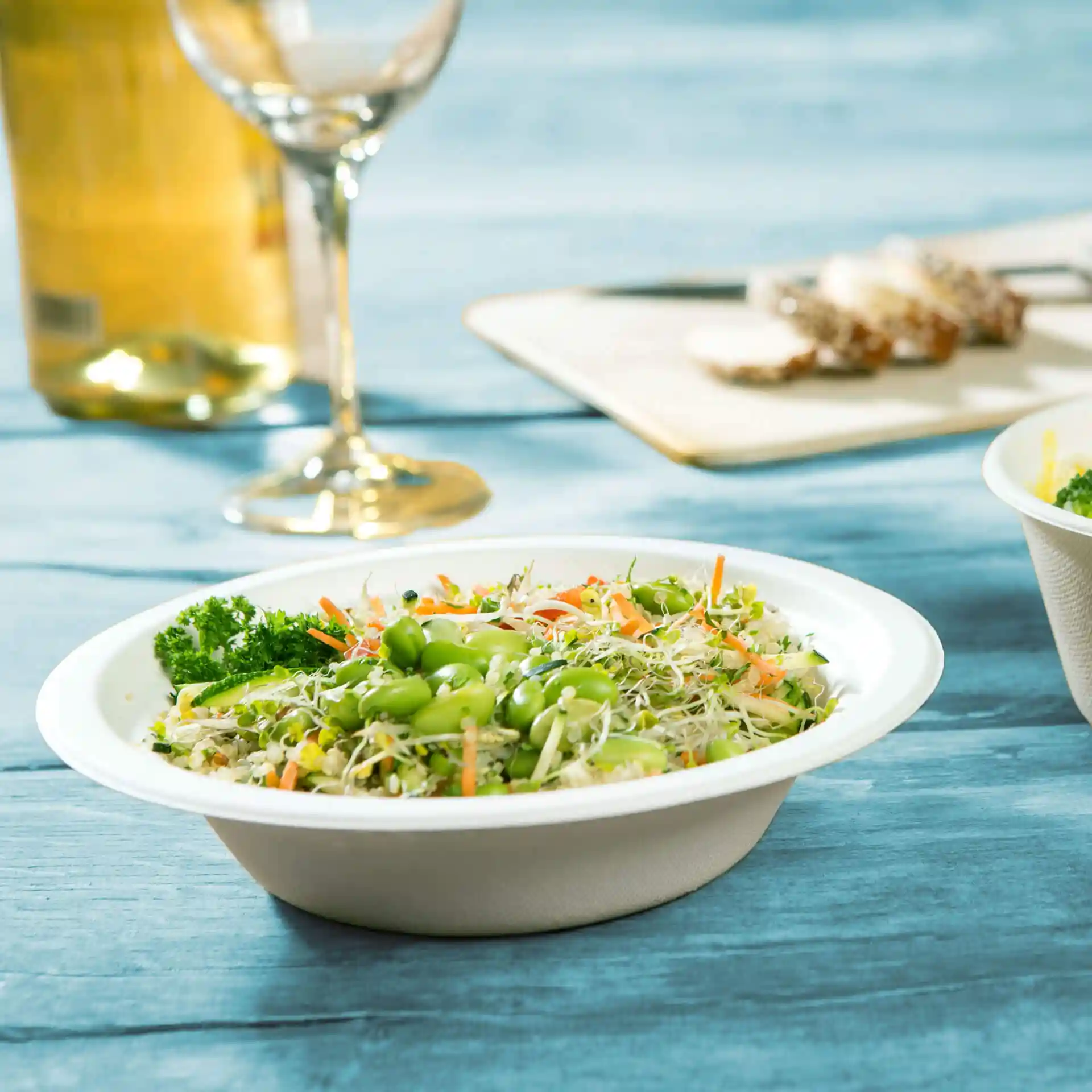 Ronde witte suikerrietkom gevuld met gemengde salade, inclusief edamame, wortelen en kiemen, op een blauwe houten ondergrond.