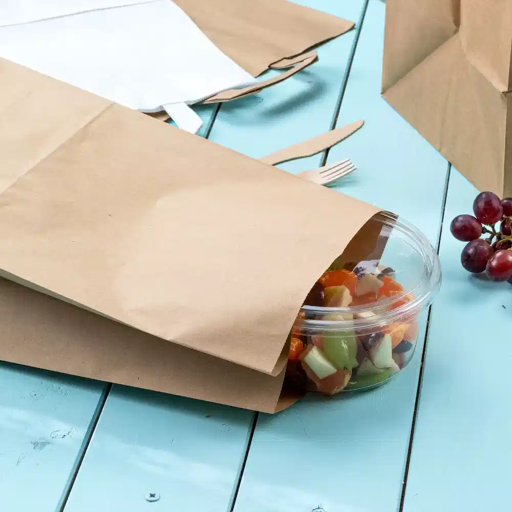 Brown papieren zakken bedekken gedeeltelijk een doorzichtige container met salade op een blauwe houten tafel. Druiven zijn in de buurt zichtbaar.