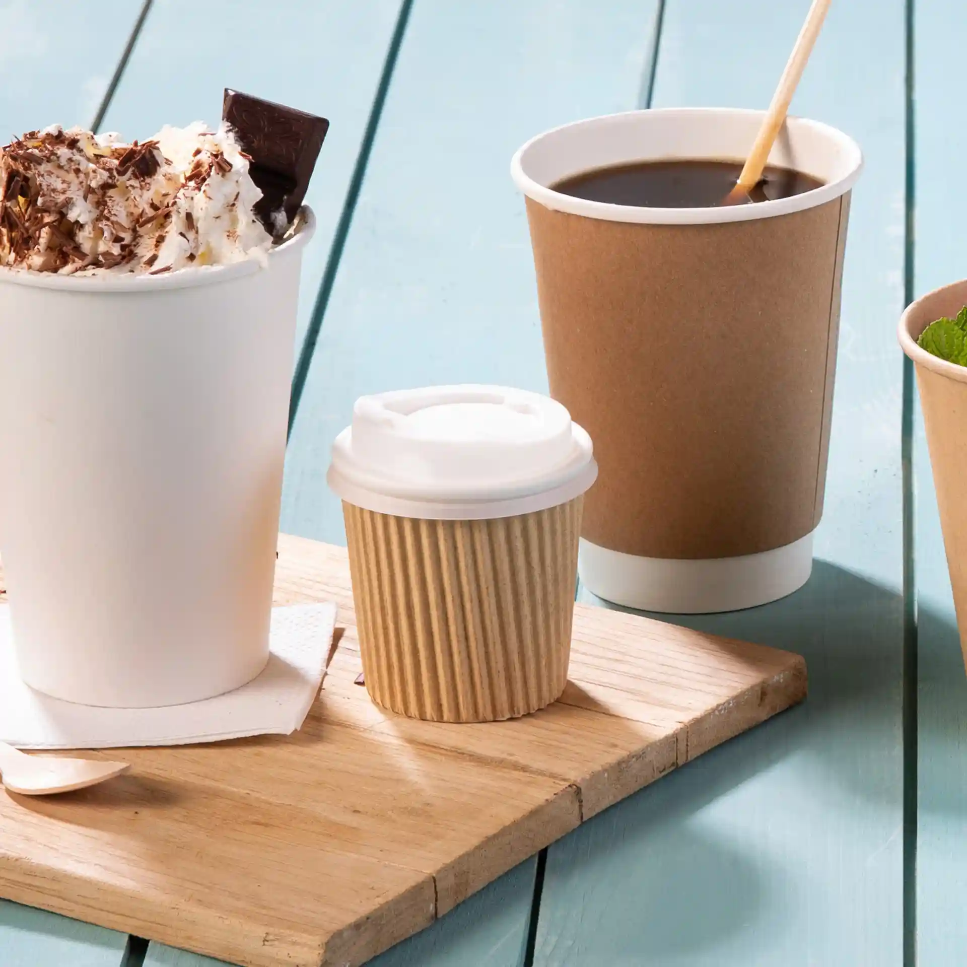 Afbeelding van een houten plank met een grote witte beker met slagroom en chocolade, een kleine ribbelige bruine beker en een bruine koffiebeker.