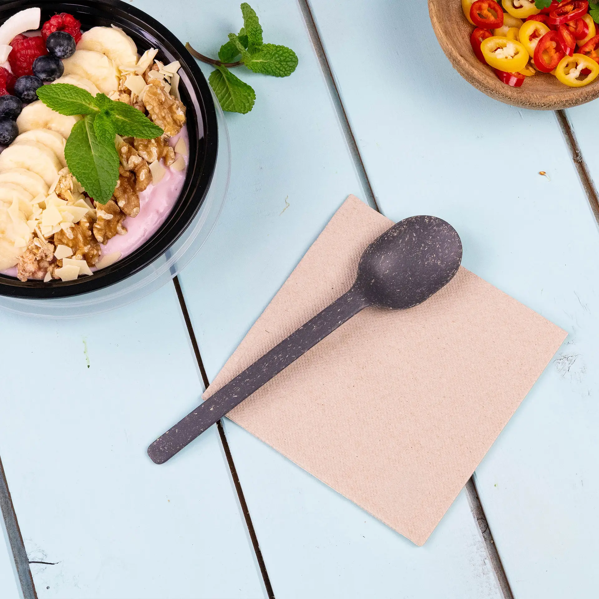 Afbeelding van een zwarte lepel op een beige servet, met een kom yoghurt met fruit en noten op de achtergrond.