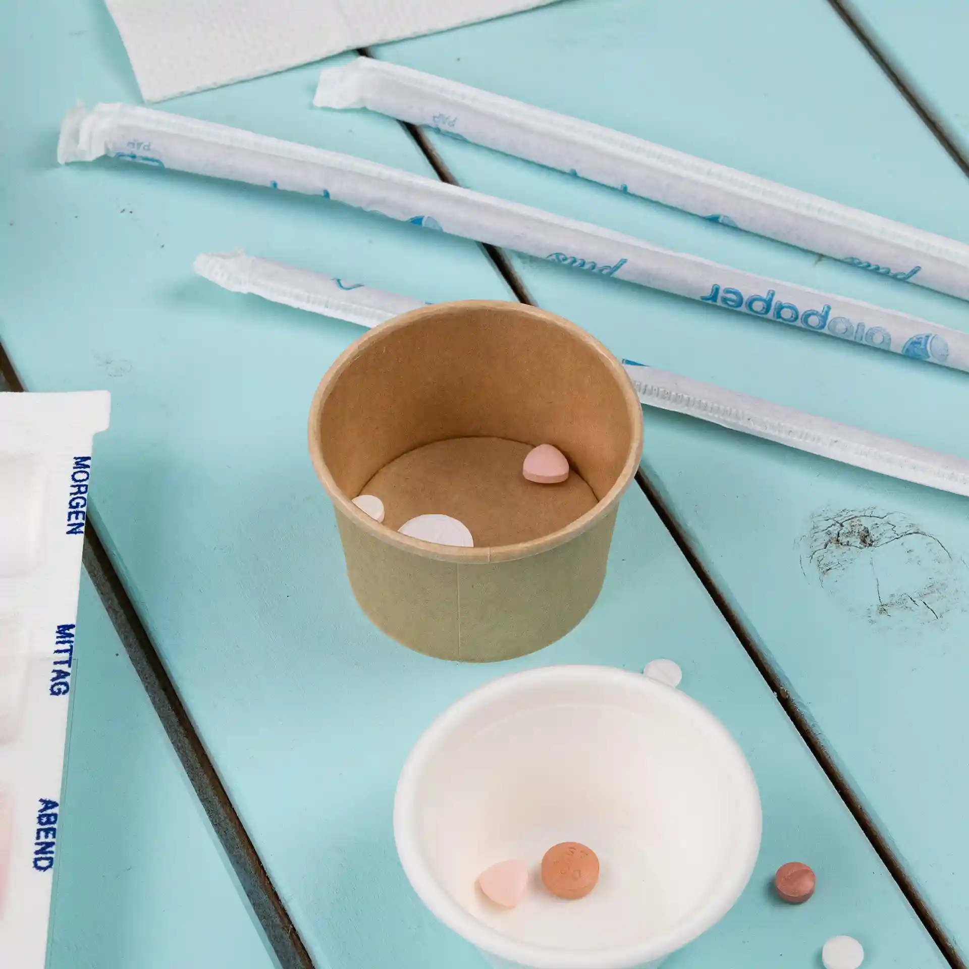 Bruine papieren beker met verschillende kleine pillen erin, geplaatst op een blauwe houten ondergrond met rietjes erbij.