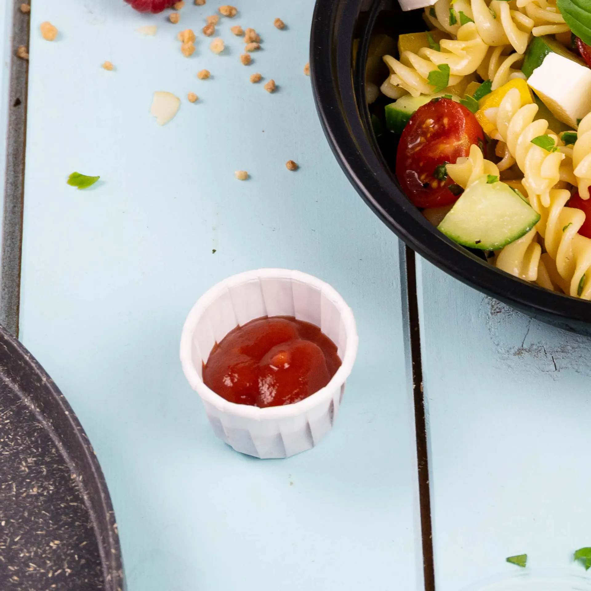 Kleine witte papieren beker gevuld met ketchup, naast een kom pastasalade op een blauwe houten ondergrond.