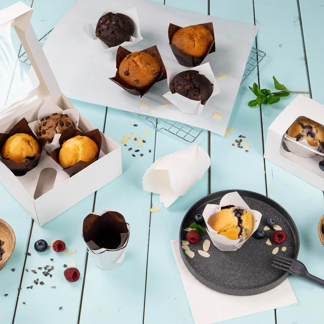 Diverse muffins in papieren wrappers op een lichtblauwe ondergrond, met een witte doos en een bord muffins en bessen.
