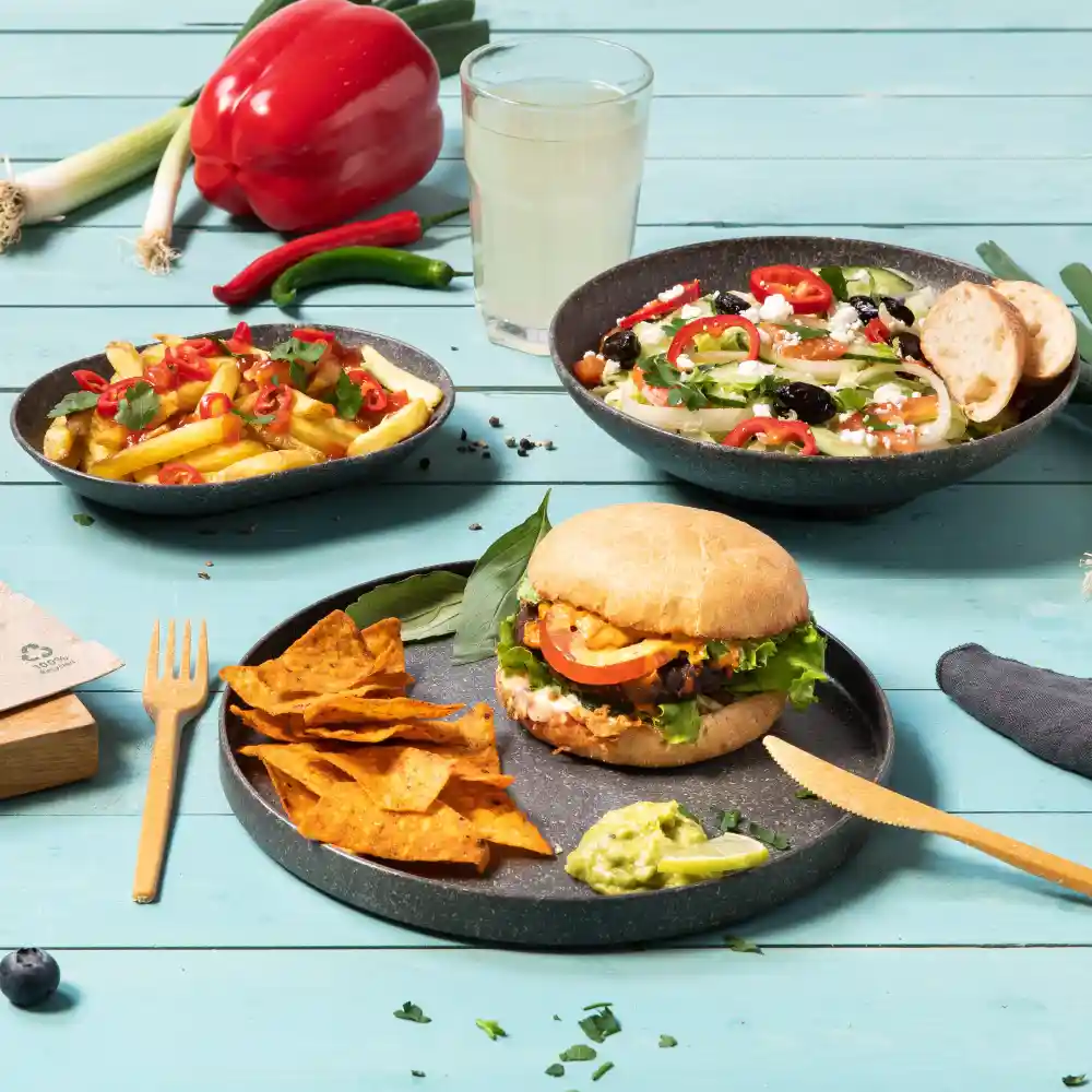 Een burger met sla, tomaat en saus, geserveerd met tortillachips en guacamole, naast een salade en friet.