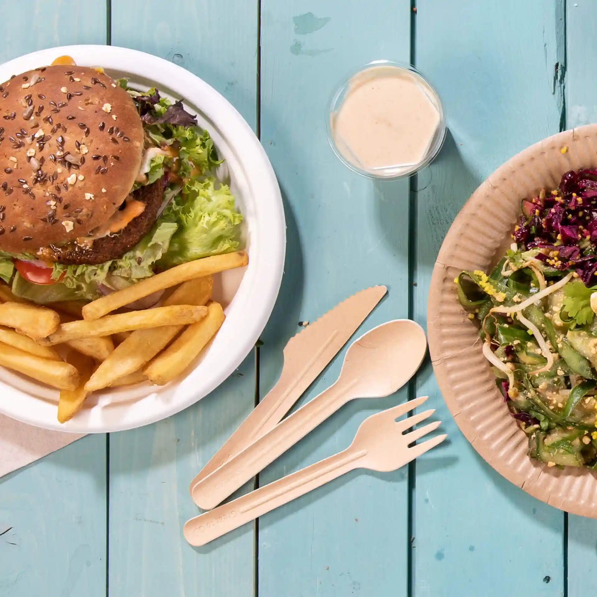 Afbeelding van een burger met sla en frietjes in een witte kom, naast een salade op een bruine papieren bord en een klein bekertje dressing.
