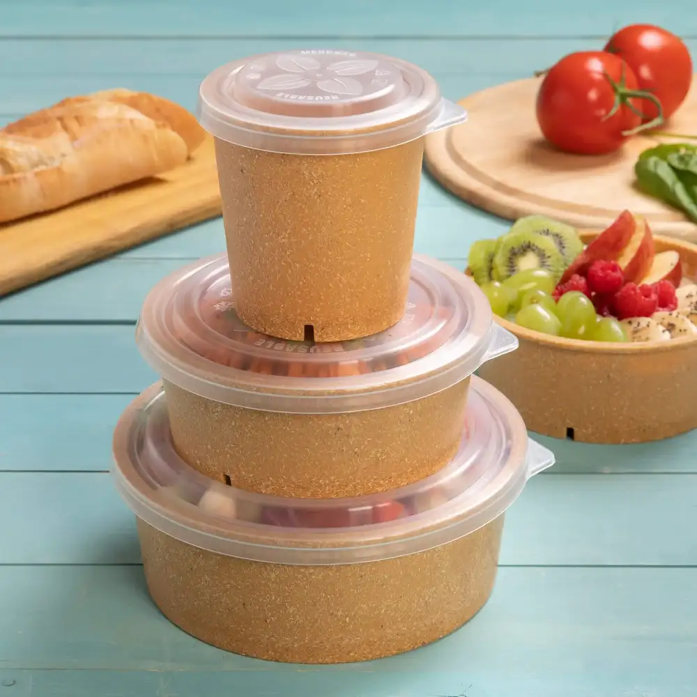 Stapel van drie bruine containers met doorzichtige deksels op een blauwe ondergrond, met brood, tomaten en fruitsalade in de buurt.