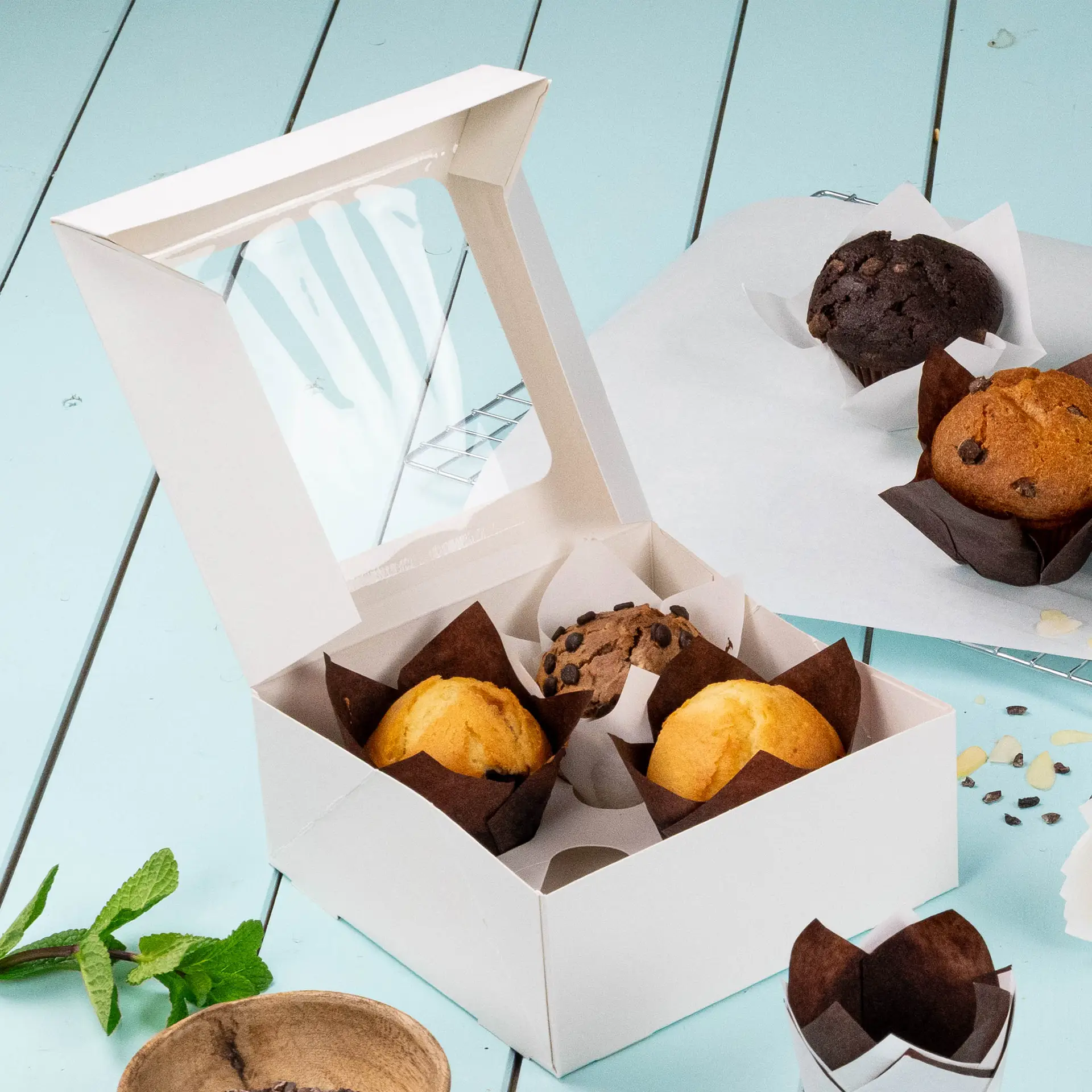 Witte cupcake-doos van 4 delen met een cellulose-venster, bevat muffins en een chocoladechipmuffin, op een lichtblauwe ondergrond.