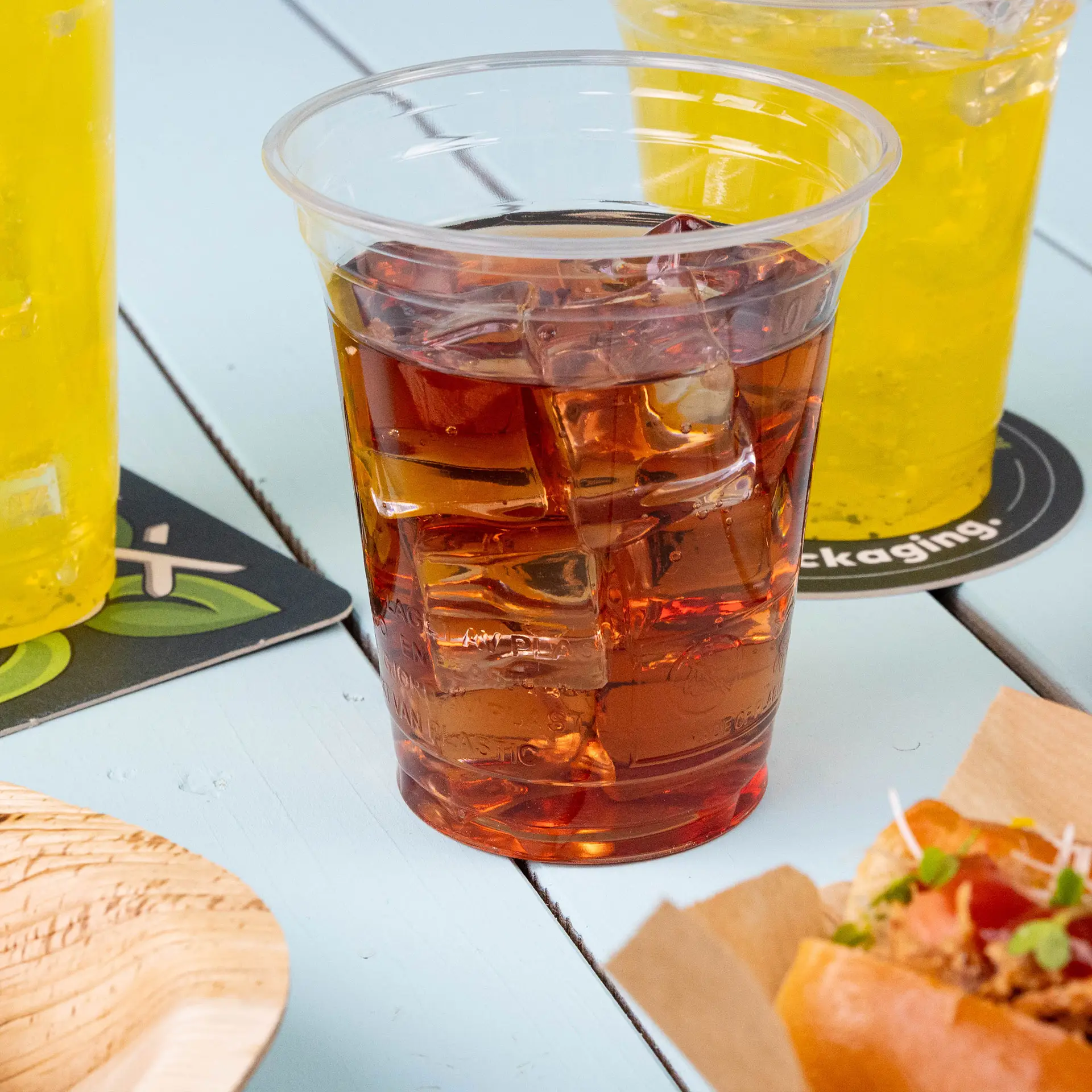 Transparante 12 oz plastic beker van PLA-bioplastic gevuld met ijsthee, omringd door gele dranken en een voedingsmiddel.